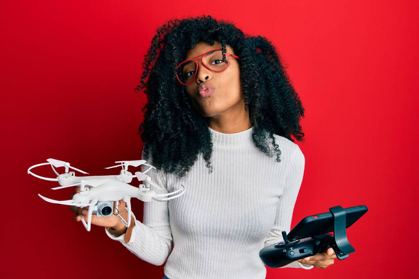 Mujer afroamericana con cabello afro usando dron con control remoto mirando a la cámara soplando un beso siendo encantadora y sexy. expresión de amor.  - Foto, Imagen