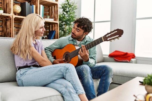 Мужчина играет на испанской гитаре со своей девушкой, сидящей дома на диване - Фото, изображение