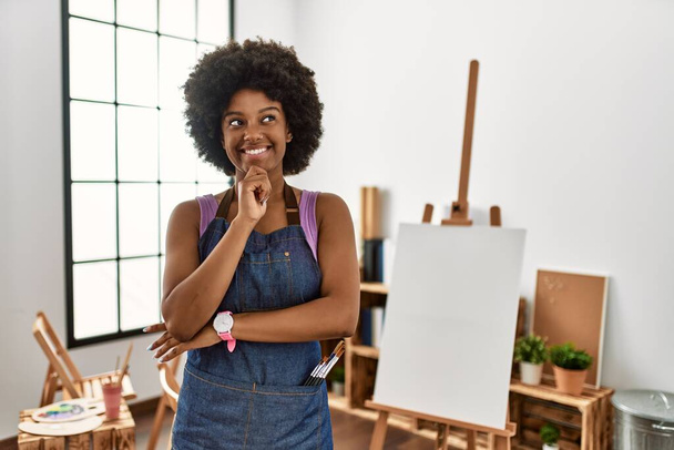 Молодая африканская американка с афро-волосами в арт-студии с рукой на подбородке думает о вопросе, задумчивом выражении лица. улыбающееся и вдумчивое лицо. Концепция сомнений.  - Фото, изображение
