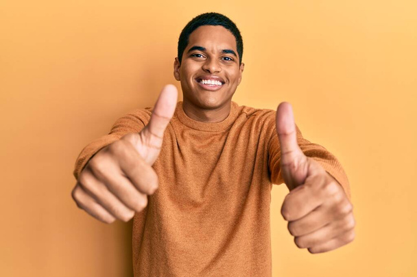 Junger gutaussehender hispanischer Mann in lässigem Winterpullover, der zustimmend positive Gesten mit der Hand macht, Daumen hoch lächelnd und glücklich über den Erfolg. Siegergeste.  - Foto, Bild