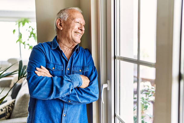 Hombre mayor con el pelo gris inclinado por la ventana de su casa, sonriendo feliz y confiado mirando afuera - Foto, Imagen