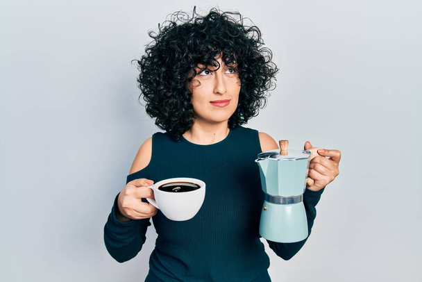 Junge Frau aus dem Mittleren Osten hält italienische Kaffeemaschine, lächelt zur Seite und starrt nachdenklich weg.  - Foto, Bild