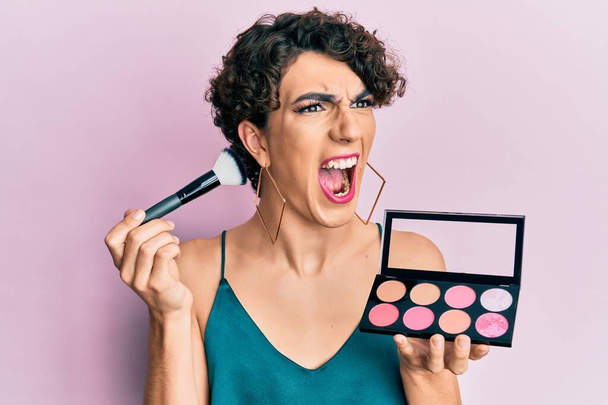 Young man wearing woman make up holding makeup brush and blush angry and mad screaming frustrated and furious, shouting with anger. rage and aggressive concept.  - Photo, Image