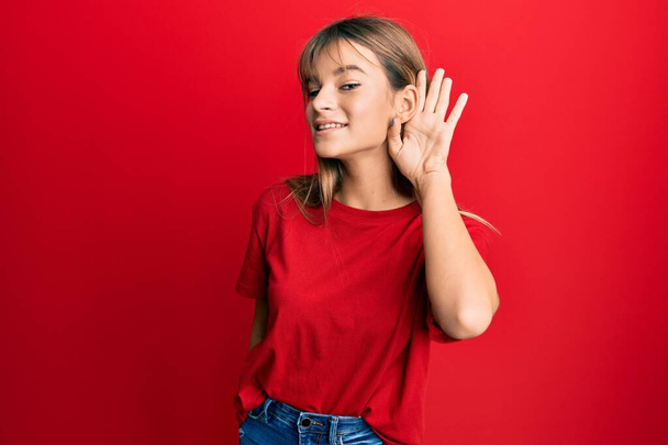 Έφηβος καυκάσιος κορίτσι φορώντας casual κόκκινο μπλουζάκι χαμογελώντας με το χέρι πάνω από το αυτί ακούγοντας και ακούγοντας φήμες ή κουτσομπολιά. έννοια της κώφωσης.  - Φωτογραφία, εικόνα