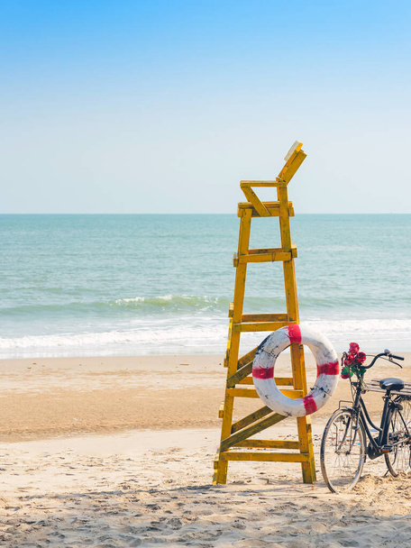 Stary retro czarny rower z bukietem kwiatów obok pierścień życia dla bezpieczeństwa życia na żółtym stanowisku ratownika lub ratownika chroniące bezpieczeństwo turystów na pustej plaży w słońcu. - Zdjęcie, obraz