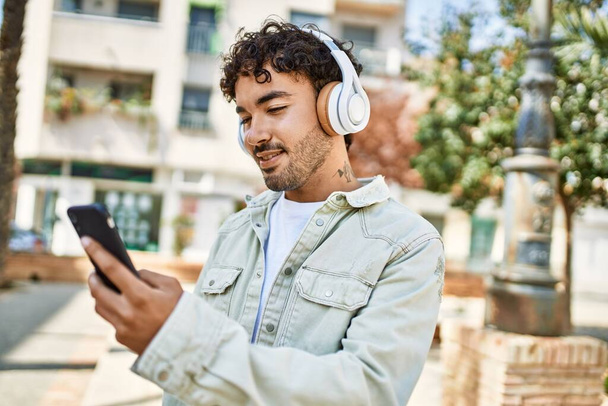 Bel homme hispanique avec la barbe souriant heureux à l'extérieur par une journée ensoleillée portant des écouteurs écoutant de la musique avec smartphone - Photo, image