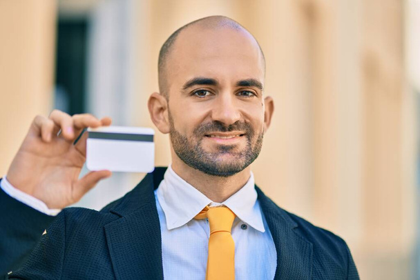 Joven empresario calvo hispano sonriendo feliz sosteniendo tarjeta de crédito en la ciudad. - Foto, Imagen