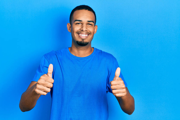 青いシャツを着た若いアフリカ系アメリカ人男性は、手で積極的なジェスチャーを行うことを承認し、親指を上げて笑顔と成功のために幸せ。勝者のジェスチャー.  - 写真・画像