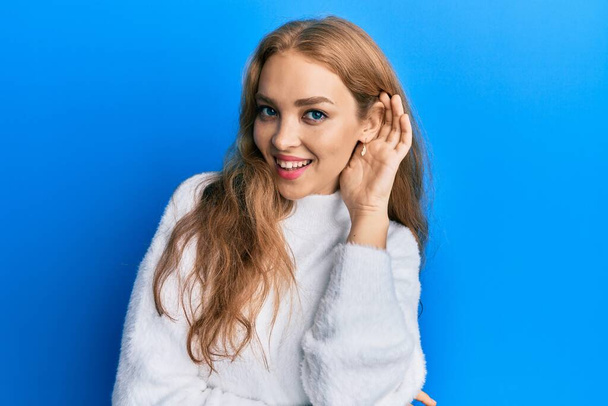 Beautiful blonde caucasian woman wearing casual winter sweater smiling with hand over ear listening and hearing to rumor or gossip. deafness concept.  - Photo, Image