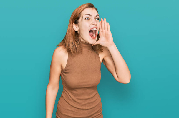 Молодая кавказская женщина в повседневной одежде кричит и громко кричит в бок с рукой на губах. коммуникационная концепция.  - Фото, изображение