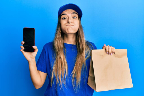 Mujer hispana joven sosteniendo bolsa de papel de entrega y mostrando la pantalla del teléfono inteligente soplando mejillas con cara divertida. boca inflada con aire, captación de aire.  - Foto, imagen