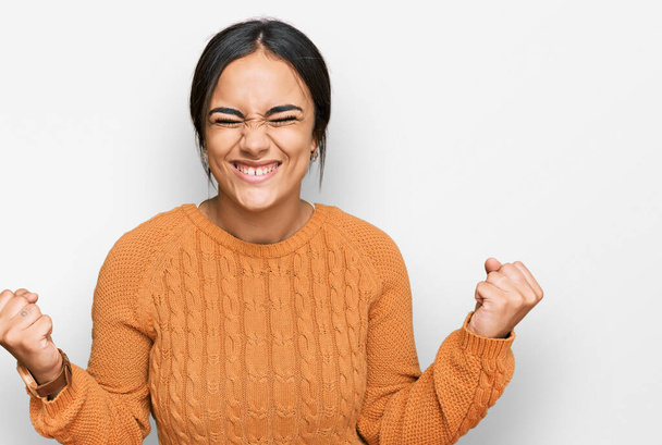 Jeune femme brune portant un pull d'hiver décontracté très heureux et excité faisant geste gagnant avec les bras levés, souriant et criant pour le succès. concept de célébration.  - Photo, image