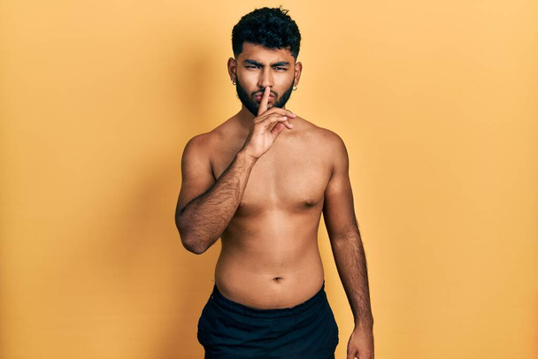Arabische man met baard draagt zwemkleding zonder shirt en vraagt om stil te zijn met de vinger op de lippen. stilte en geheim concept.  - Foto, afbeelding