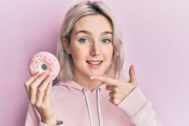 Young blonde girl holding donut smiling happy pointing with hand and finger  - Foto, Imagem