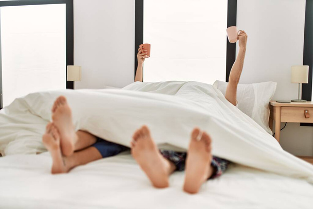 Un paio svegli sotto coperta. Facendo colazione tenendo una tazza di caffè sollevata sul letto. - Foto, immagini