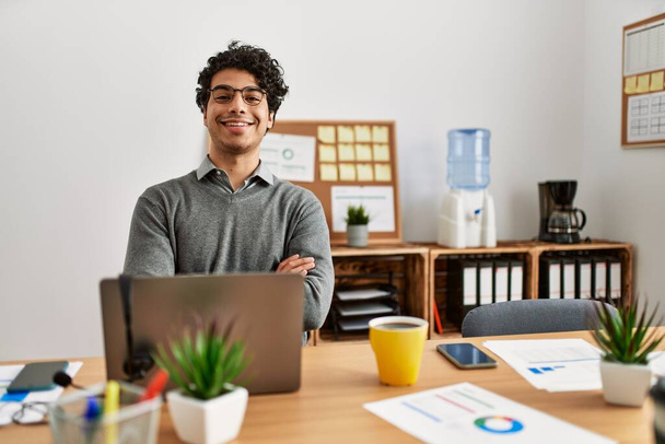 Joven hombre hispano con estilo de negocios sentado en el escritorio en la oficina cara feliz sonriendo con los brazos cruzados mirando a la cámara. persona positiva.  - Foto, imagen