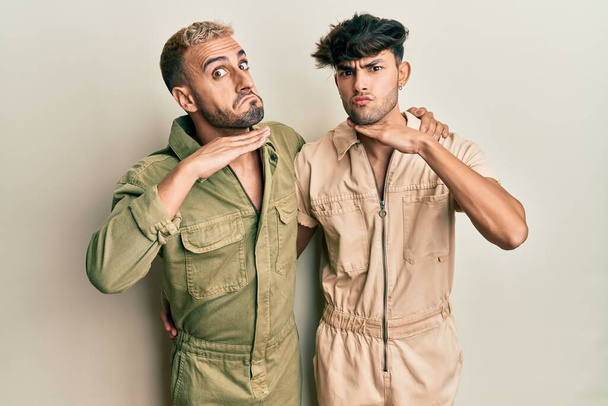 Homosexual gay couple standing together wearing casual jumpsuit cutting throat with hand as knife, threaten aggression with furious violence  - Photo, Image