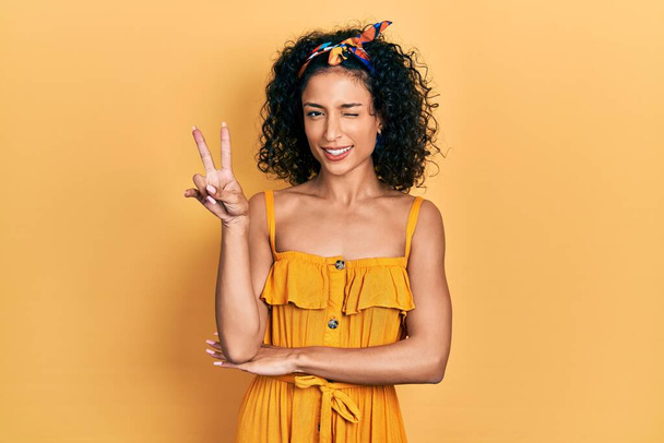 Young latin girl wearing summer dress smiling with happy face winking at the camera doing victory sign. number two.  - Photo, Image