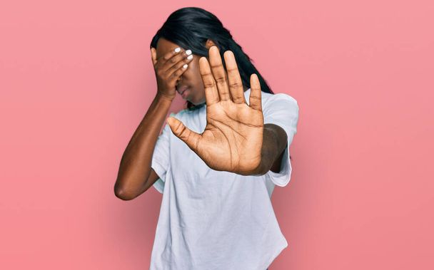 Молода афроамериканська жінка, одягнена в білу сорочку, закриває очі руками і припиняє жести з сумним і страхом. соромливе і негативне поняття.  - Фото, зображення