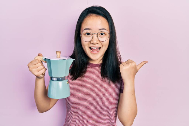 Молодая китайская девушка держит итальянскую кофеварку указывая пальцем вверх в сторону улыбается счастливо с открытым ртом  - Фото, изображение