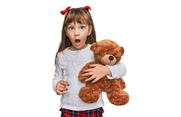 Little caucasian girl kid hugging teddy bear stuffed animal scared and amazed with open mouth for surprise, disbelief face  - Photo, Image