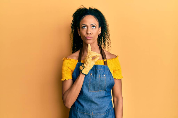 Middle age african american woman wearing professional apron thinking concentrated about doubt with finger on chin and looking up wondering  - Fotografie, Obrázek