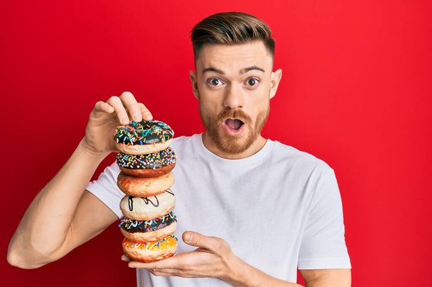 Junger rothaariger Mann mit einem Haufen leckerer bunter Donuts, verängstigt und schockiert von Überraschung und erstaunter Miene, Angst und aufgeregtem Gesicht.  - Foto, Bild