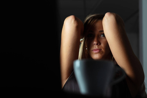 Zmęczona kobieta pracuje do późna przy laptopie w ciemnym biurze, podnosząc ręce do głowy, patrząc przed kamerę. - Zdjęcie, obraz