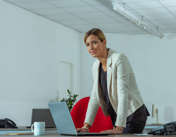 Smiling confident businesswoman standing over her laptop computer in a spacious modern office in a low angle view with copyspace - Zdjęcie, obraz