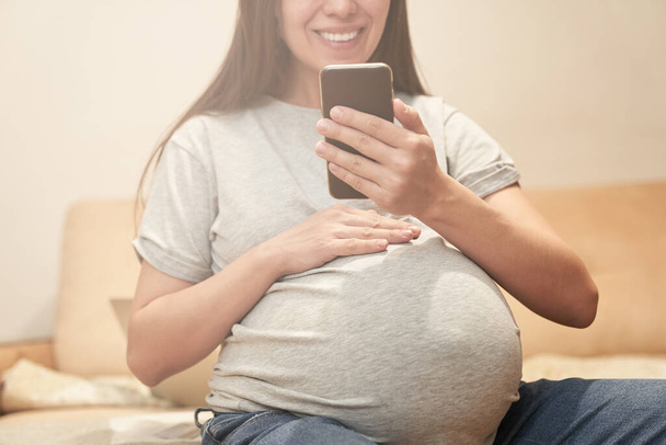 Raskaana oleva nainen kotona puhelimen kanssa istuu ja hymyilee, videoviestintä - Valokuva, kuva