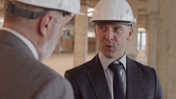 Poitrine du superviseur de construction caucasien masculin portant une combinaison formelle et un casque blanc, parlant à un collègue sur le chantier - Séquence, vidéo