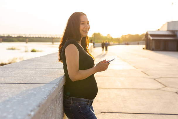 Femme enceinte avec smartphone à l'extérieur. Grossesse, technologie et concept de communication - Photo, image