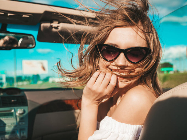 Porträt einer jungen schönen und lächelnden Hipsterfrau im Cabrio. Sexy unbeschwerte Cabrio-Fahrerin. Positives Modellfahren und Spaß mit Sonnenbrille bei Sonnenuntergang - Foto, Bild