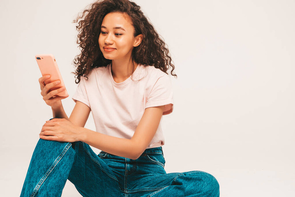 Beautiful black woman with afro curls hairstyle.Smiling model in white trendy jeans clothes. Sexy carefree female sitting on grey background in studio. Holding smartphone in hands. Using apps  - Φωτογραφία, εικόνα