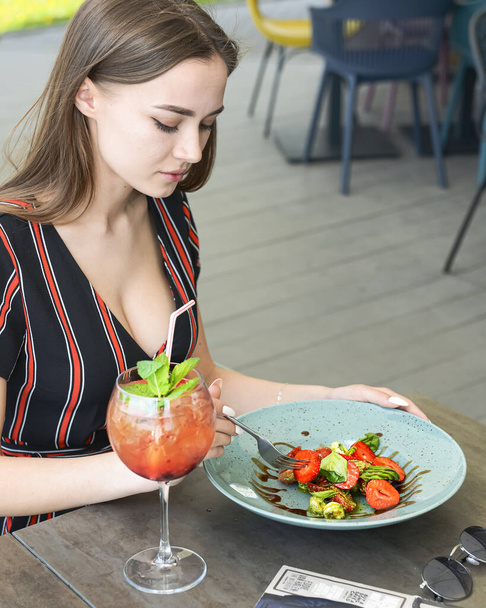 Young woman eating mixed caprese salad with strawberries and drinking aperol spritz soda cocktail in outdoor restaurant. Eat out concept, eating alone due to worldwide pandemic. - Фото, изображение