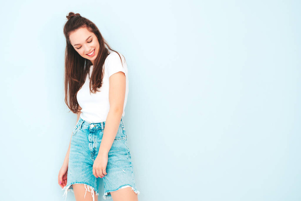Young beautiful smiling female in trendy summer hipster clothes. Sexy carefree woman posing near light blue wall in studio. Positive model having fun indoors. Cheerful and happy - 写真・画像