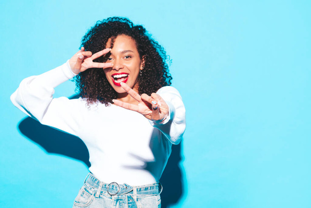 Beautiful black woman with afro curls hairstyle.Smiling model in trendy summer clothes. Sexy carefree female posing near blue wall in studio.With red lips.Shows peace sign - Foto, immagini