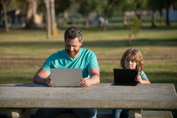 Daddy and son sitting at park and educating on laptop. Family education Concept. Father teaching his little toddler boy how to use personal computer, laptop. - 写真・画像