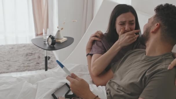Mediana PAN inyección de feliz pareja latina con prueba de embarazo sentado en la cama. Emocional joven llorando mientras su marido la abraza compartiendo este momento íntimo juntos en casa - Metraje, vídeo