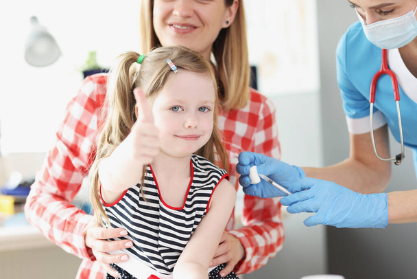 Petite fille tient ses pouces vers le haut et le médecin vaccine son épaule dans le bureau de médina - Photo, image