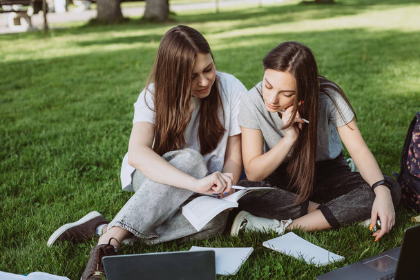 Две студентки сидят в парке на траве с книгами и ноутбуками, учатся и готовятся к экзаменам. Дистанционное образование. Мягкая селективная фокусировка. - Фото, изображение