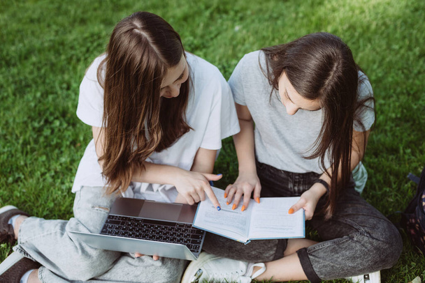 Due studentesse sono sedute nel parco sull'erba con libri e computer portatili, studiano e si preparano per gli esami. Istruzione a distanza. Concentrazione selettiva morbida. - Foto, immagini