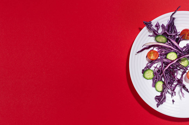 Fitness fresh salad of raw vegetable with purple cabbage on half white plate in sun light with shadow on red color background, top view. - Fotó, kép