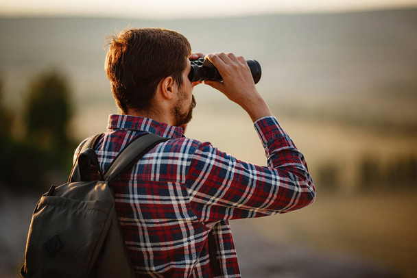 丘の双眼鏡を見ている男。バックパック付きのTシャツの男。谷の風景の中にハイキング中の若い白人男性 - 写真・画像