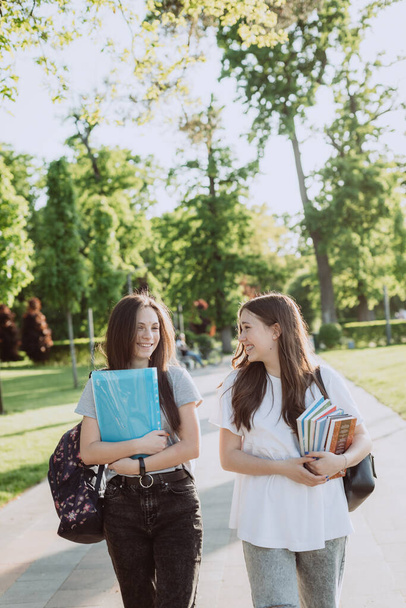 2人の幸せな笑顔の学生の女の子が歩いていて、暖かい晴れた日にキャンパスでお互いに話しています。柔らかい選択的フォーカス. - 写真・画像