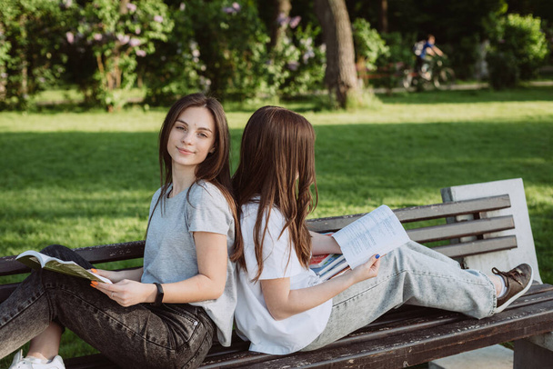 Two student girls are looking at an open book on a bench in the park. Distance education, preparation for exams. Soft selective focus. - Photo, Image