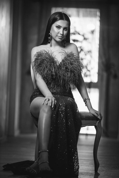 A vertical greyscale shot of a young brunette female posing sitting on a chair in a evening dress - Foto, Imagen