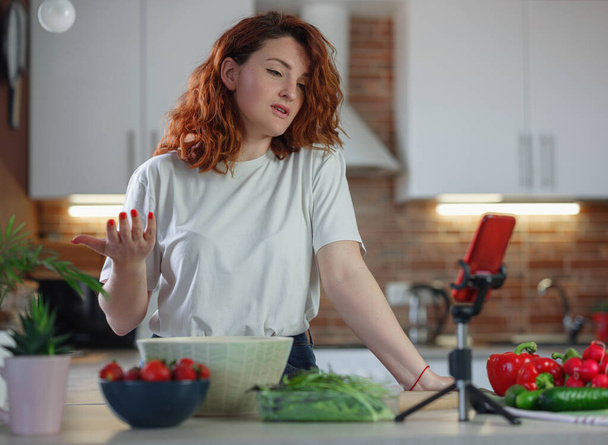 Young pretty woman blogger shoots a video of a salad recipe on a smartphone camera. The concept of healthy eating. Food blog concept - Photo, Image