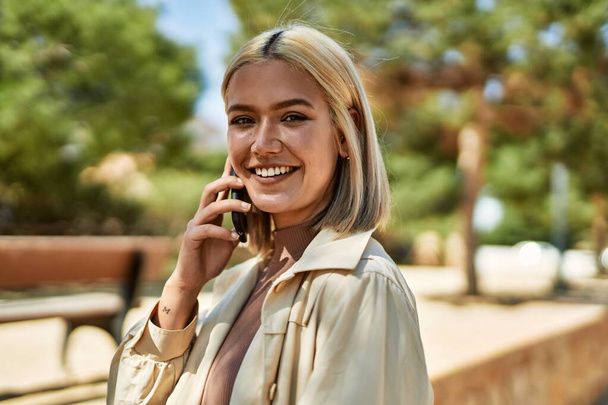 Молодая блондинка улыбается счастливо разговаривая по смартфону в городе. - Фото, изображение