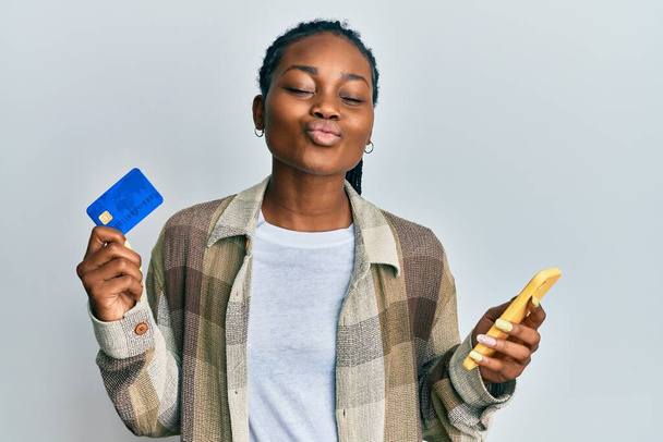 Joven mujer afroamericana sosteniendo teléfono inteligente y tarjeta de crédito mirando a la cámara soplando un beso siendo encantador y sexy. expresión de amor.  - Foto, Imagen